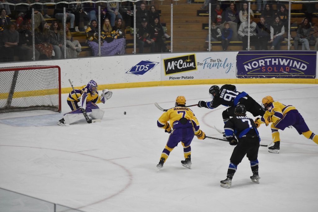 Hockeyottelussa mustapaitainen pelaaja laukoo kiekon maalia kohti, kun neljä vastustajaa puolustaa alhaalla.