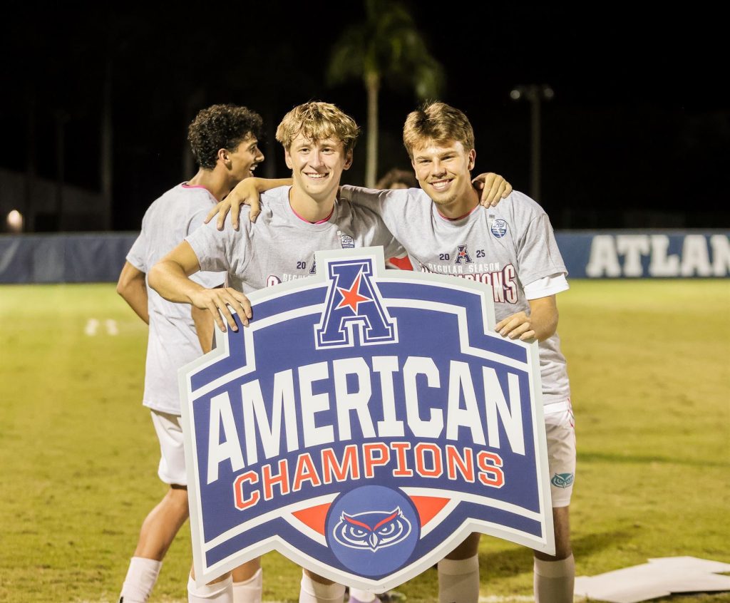 Kaksi nuorta miestä seisoo jalkapallokentällä pitäen yllä suurta julistetta, jossa lukee "AMERICAN CHAMPIONS", taustalla on ilta-ajan tunnelma.